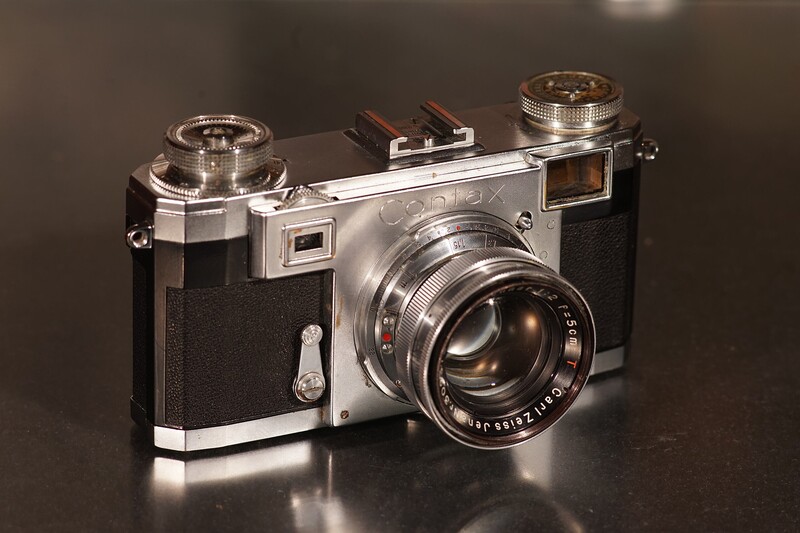 Vintage rangefinder camera with chrome and black body, bright lens, and dual viewfinders against dark background.