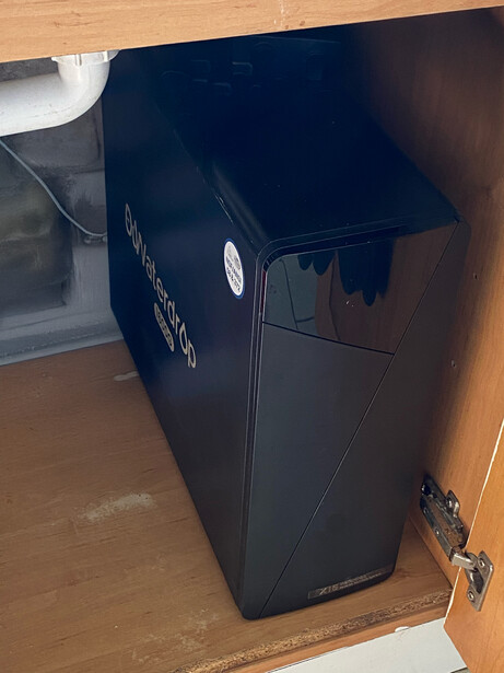 Black UPS uninterruptible power supply unit mounted under a wooden shelf indoors.