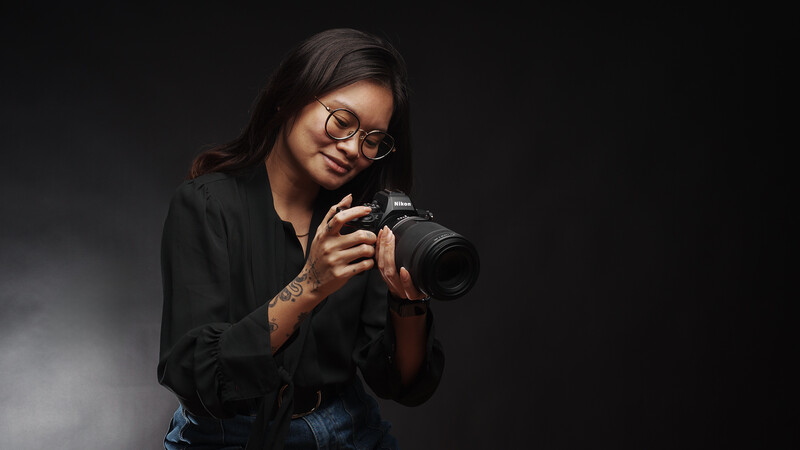 Photographer with glasses holding a DSLR camera with telephoto lens, smiling at camera against dark background.
