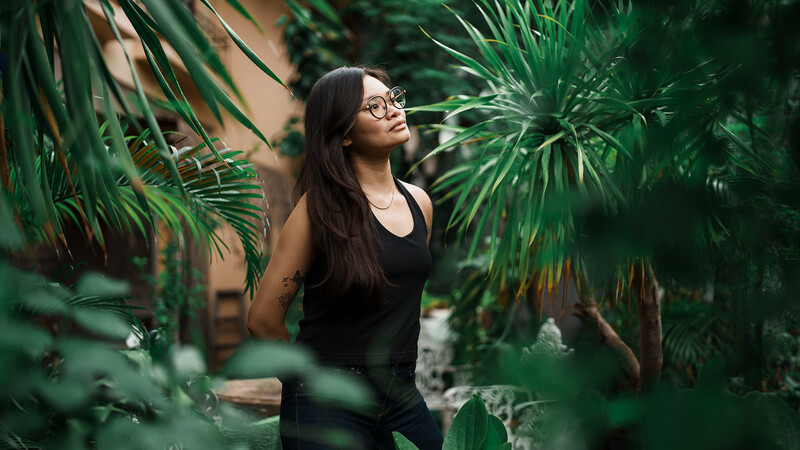 Woman in black tank top and glasses surrounded by lush green foliage, looking upward thoughtfully.
