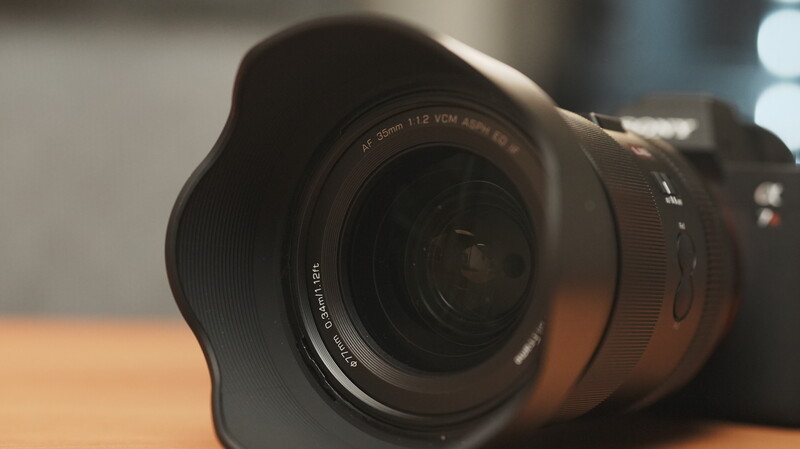 Wide-angle lens with hood attached, positioned on wooden surface with soft directional lighting.