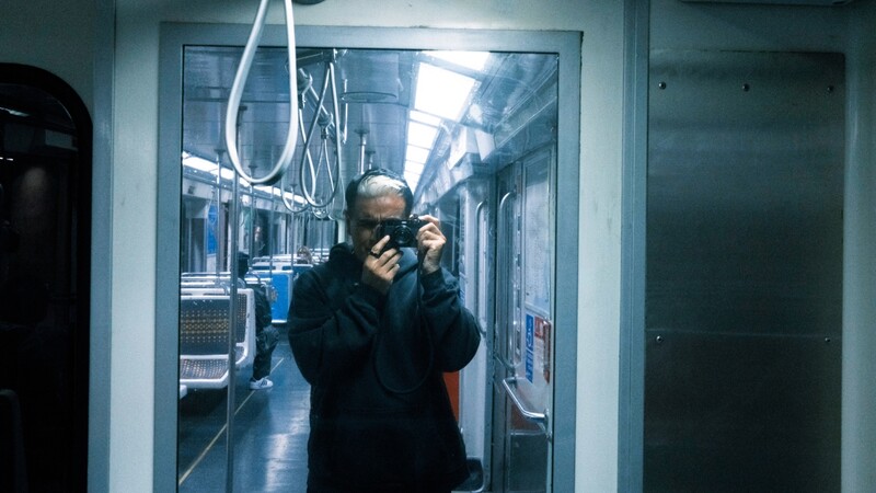 Man in dark jacket holding camera inside modern subway car with overhead cable visible.