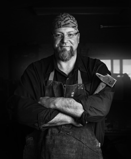 Bearded man wearing a bandana and work apron, arms crossed, in a black and white portrait.