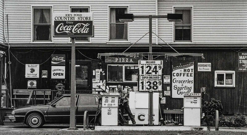 Black and white photograph of a vintage country store front with gas pump, vintage signage, and parked car.