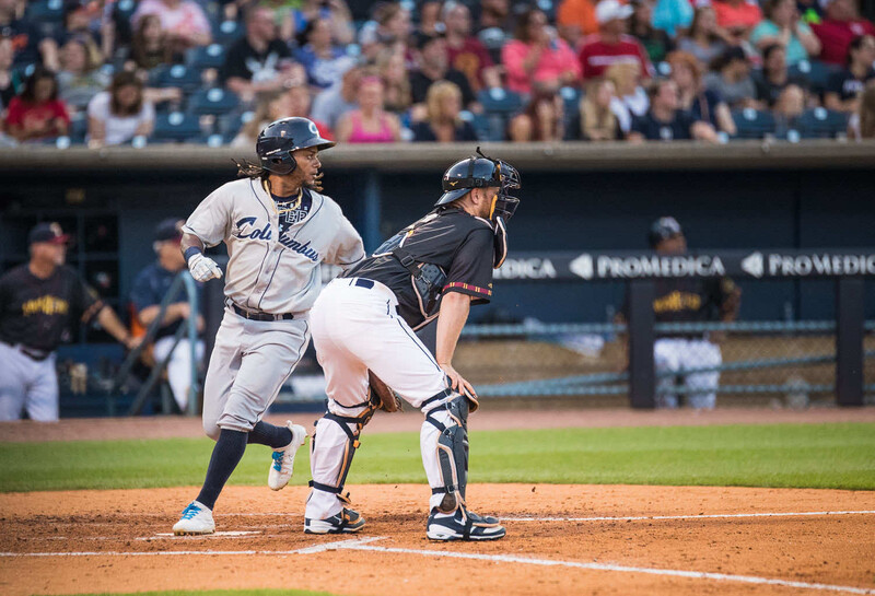 Baseball player running to first base while catcher crouches during game with crowded stadium in background.