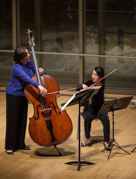 Cellist in blue shirt performs with violinist in black at wooden stage with large windows.
