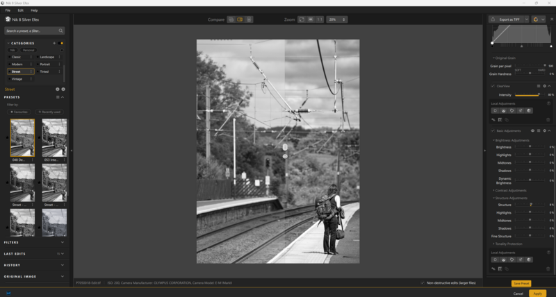 Black and white photograph of a person standing on a train platform with power lines and hillside visible.