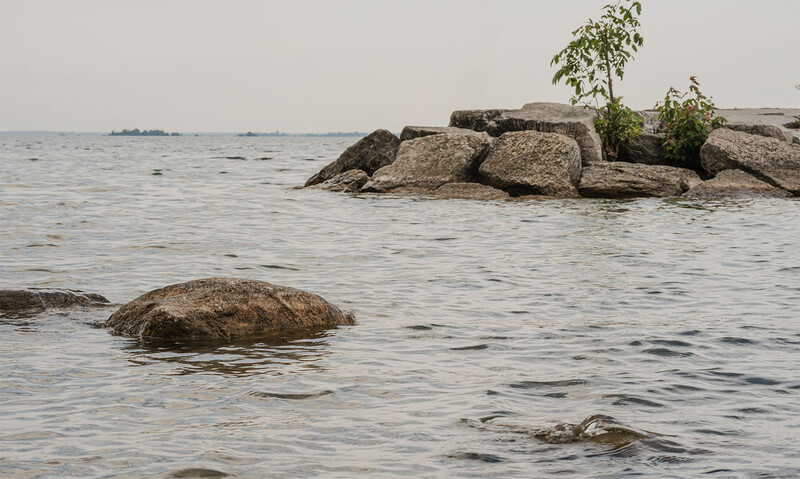 Rocky shoreline with boulders in calm water and sparse vegetation on distant rock formation.