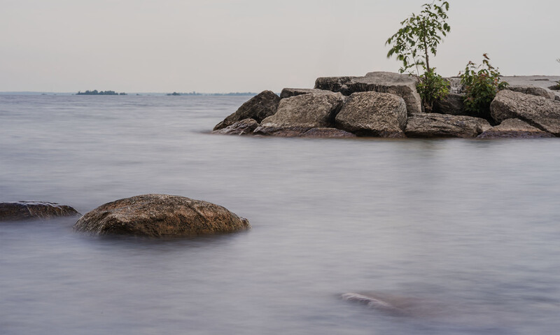 Smooth water surrounding scattered boulders with vegetation, creating a serene coastal or lakeside scene.