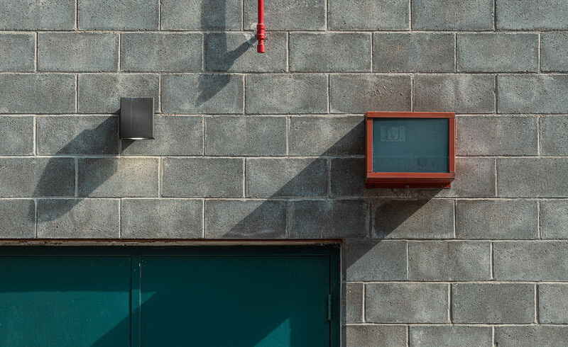 Minimalist concrete block wall with red-framed window, dark door, and cast shadows in bright sunlight.