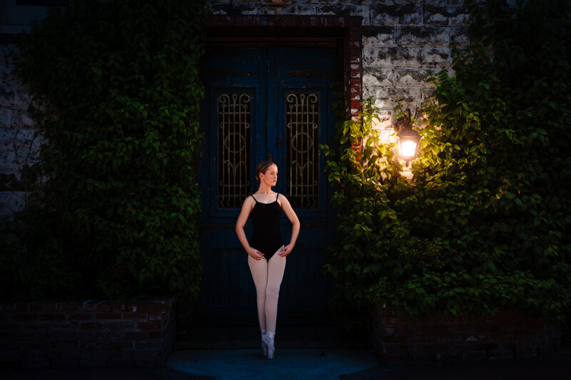 Ballerina in black leotard and pointe shoes posed in doorway framed by ivy vines and architectural details.