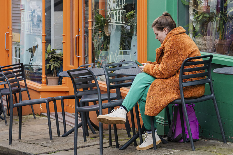Woman in orange coat and turquoise pants sitting at outdoor café table with purple bag.