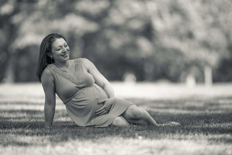 Pregnant woman in light dress seated on grass, smiling toward camera in outdoor maternity portrait.