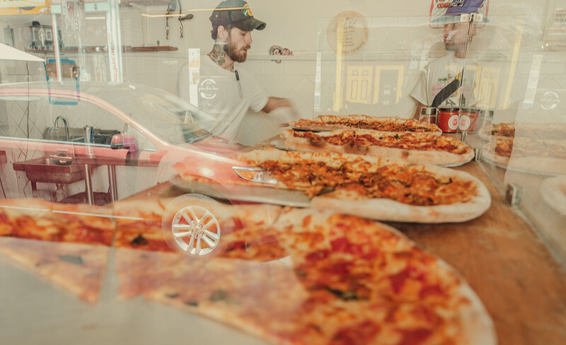 Multiple pizzas with toppings arranged on counter in commercial kitchen with workers in background.