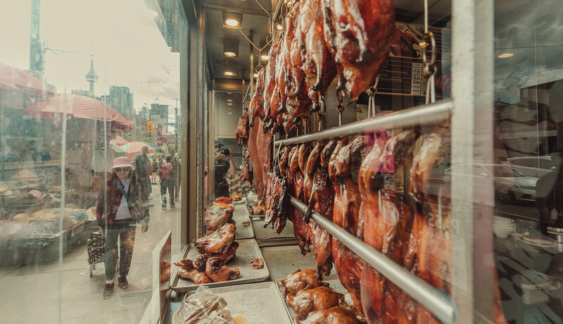 Roasted ducks hanging in a shop window with a city street and pedestrians visible through the glass.