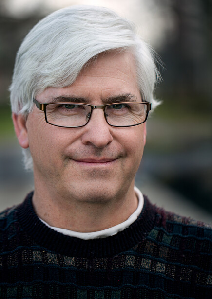 Portrait of a man with white hair and glasses wearing a dark shirt against a blurred outdoor background.