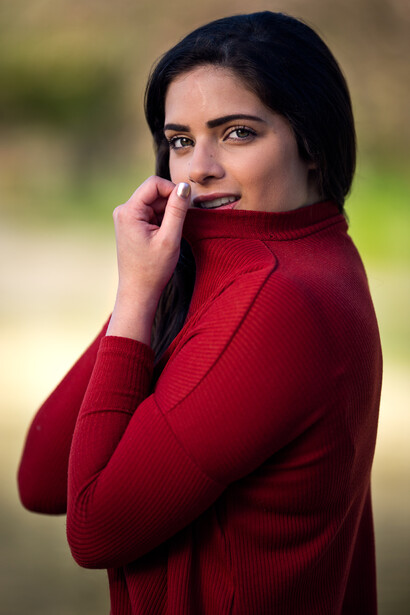 Woman in red sweater with hand near face, looking at camera with neutral expression.