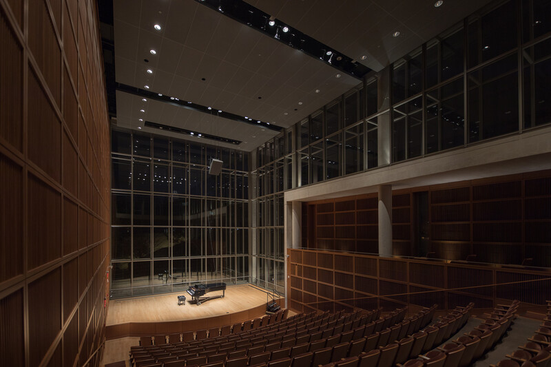 Modern auditorium with wooden wall panels, floor-to-ceiling glass windows, stage with lighting, and tiered seating.
