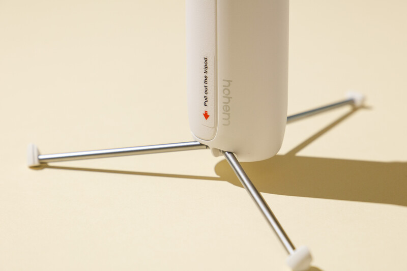 Minimalist product photography of a white cylindrical device with metal tripod legs casting shadows on a neutral beige background.