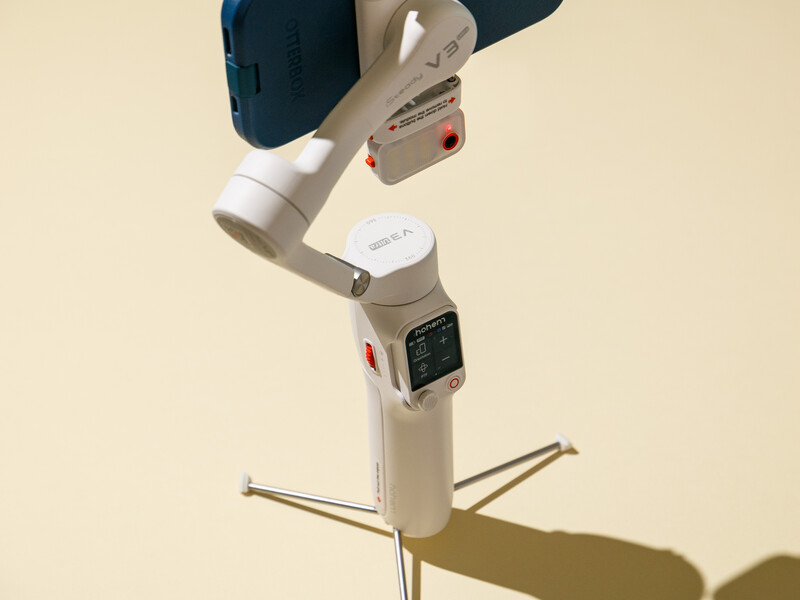 White gimbal stabilizer with articulating arms mounted on tripod against cream background.