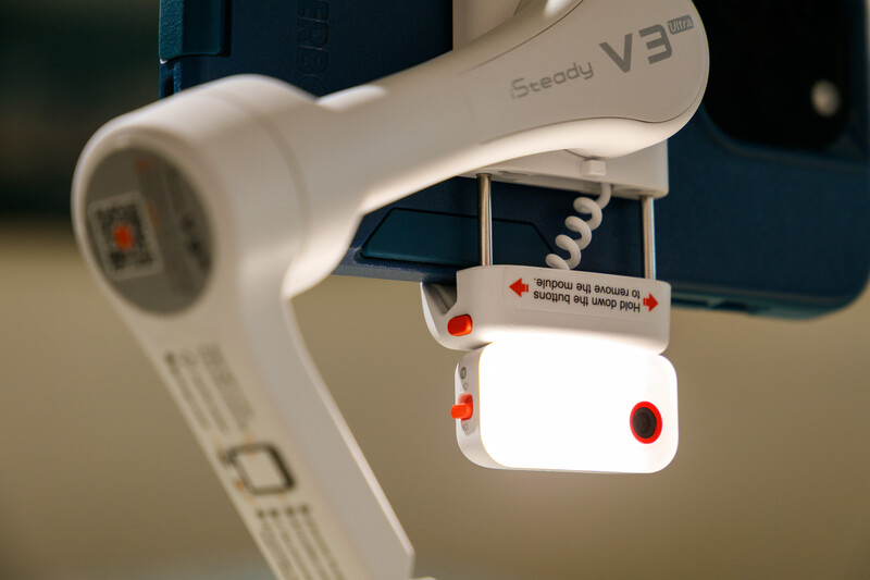White gimbal stabilizer with suspended wireless microphone and red accents against blurred background.