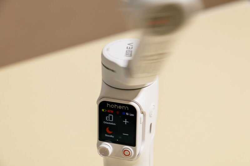 White gimbal stabilizer with control panel featuring digital display and buttons mounted on pale surface.