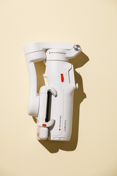 White and gray handheld device with strap and red indicator dot, photographed flat-lay against cream background.