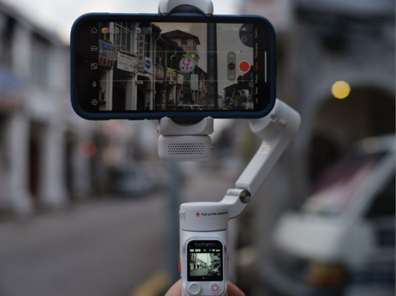 Gimbal-stabilized smartphone mounted on a three-axis stabilizer displaying a street scene through its camera app.