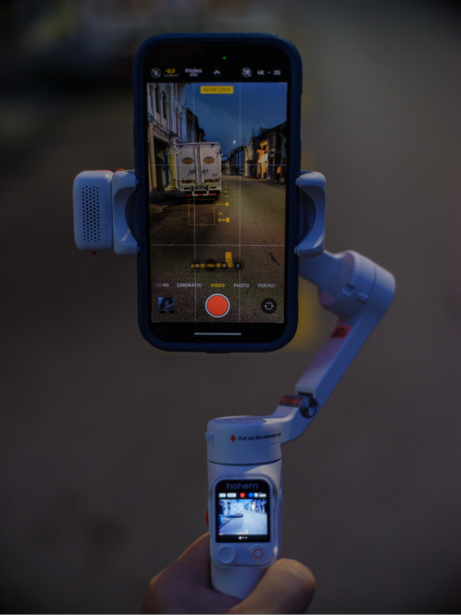 Handheld gimbal stabilizer with attached smartphone displaying video recording interface of an urban street scene.