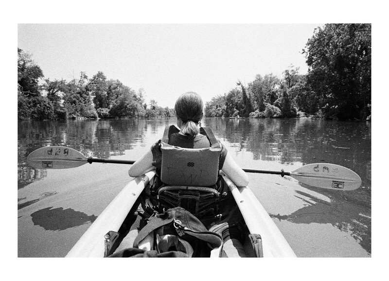 Person paddling a kayak down a calm river lined with dense trees, shot from behind in black and white.