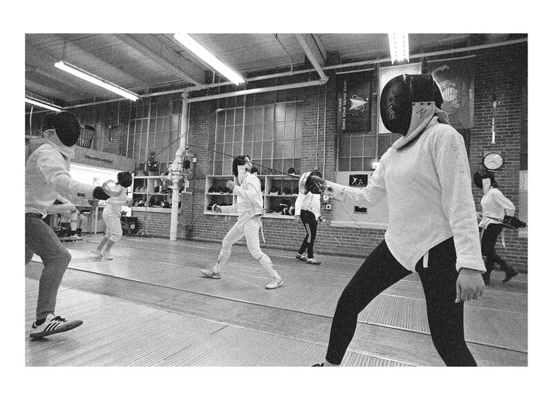 Fencers in white uniforms practicing in an indoor gym with protective masks and foils.