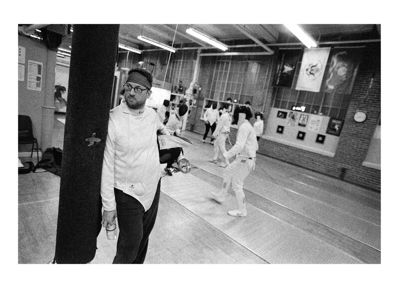 Man in white shirt and glasses holds camera while standing in gymnasium with other people in background.
