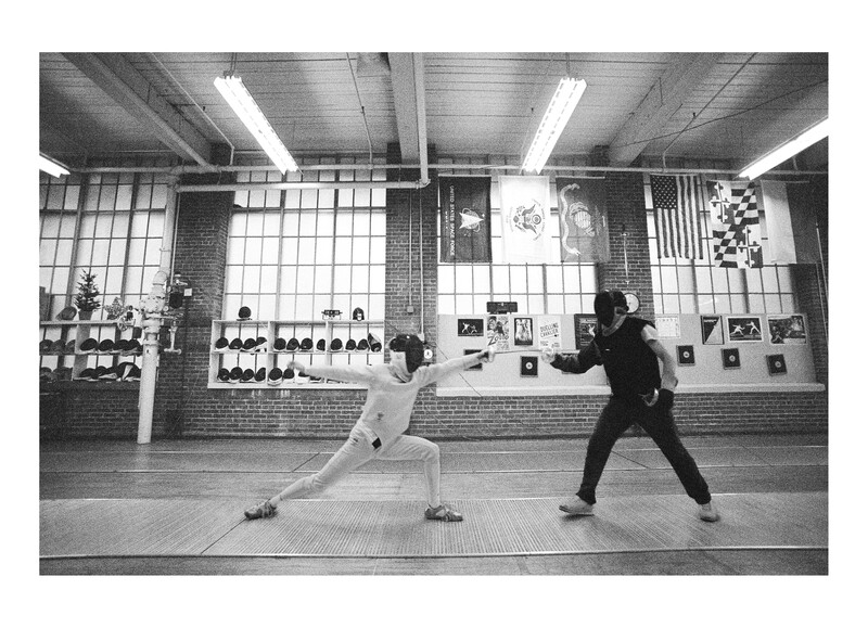 Two fencers in a gymnasium engage in a match, one in white and one in black, with weapons raised.