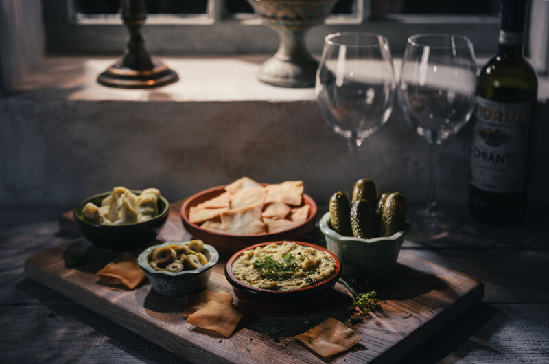 Styled flat lay of appetizers, wine, and glassware arranged on a wooden board with dramatic shadows.