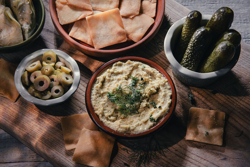 Overhead arrangement of Mediterranean appetizers including hummus, cured meats, olives, pickles, and pita on wooden table.
