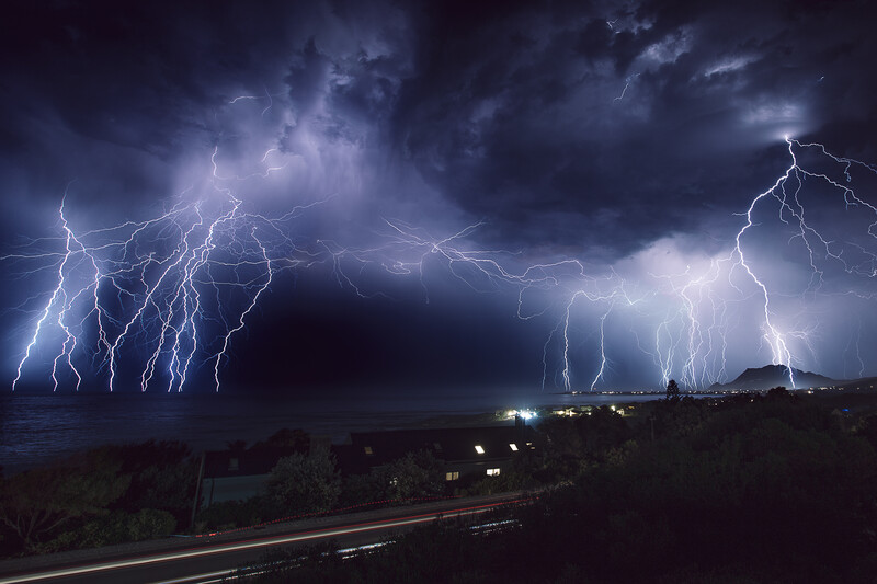 Intense lightning storm with multiple branching bolts illuminating dark clouds above a distant coastline at night.