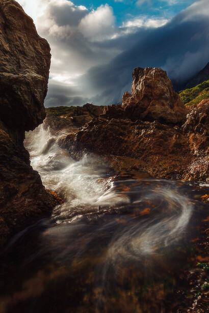 Swirling ocean waves crash against rocky coastal cliffs beneath dramatic clouds.
