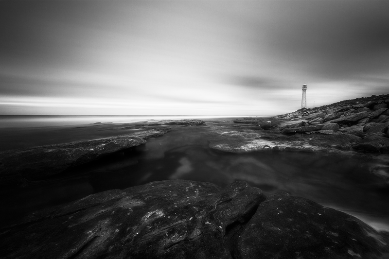 Rocky coastal landscape with a distant lighthouse under dramatic cloudy skies.