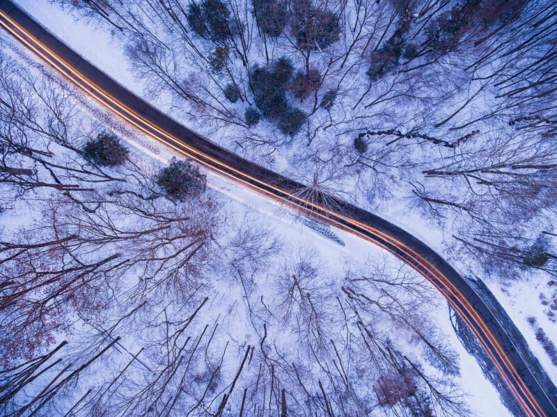 Aerial view of a curved road through snow-covered forest with bare trees and vehicle light trails.