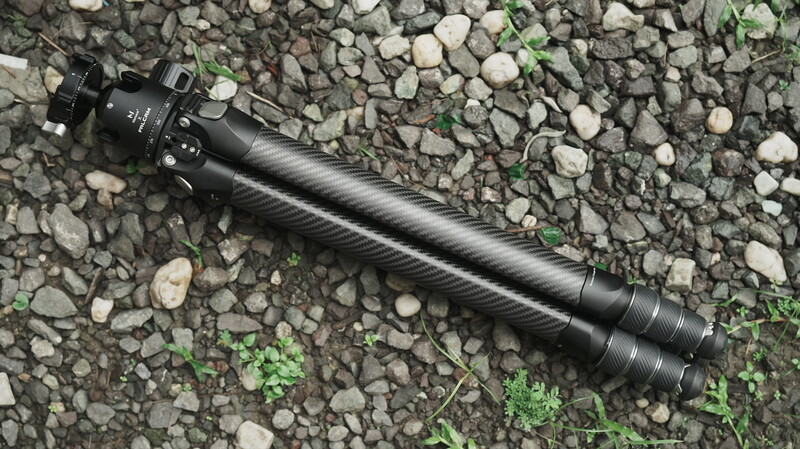 Black carbon fiber rifle suppressor with tactical mounting hardware on gravel and moss ground.