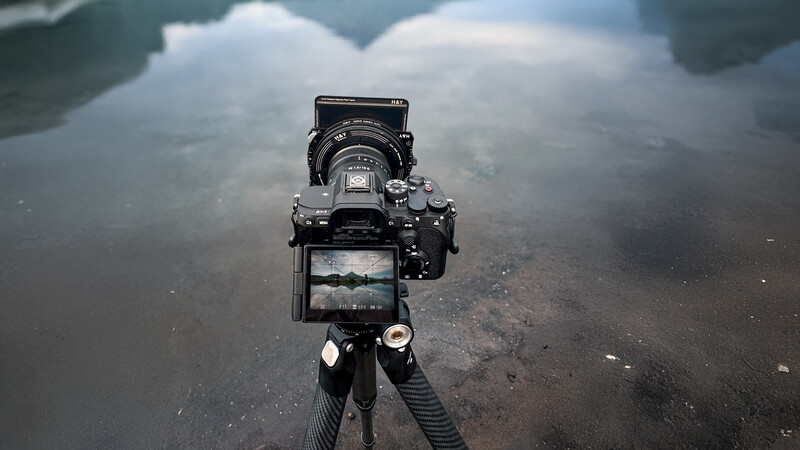 Professional mirrorless camera on tripod with articulating monitor displaying live view of cloudy sky and water reflection.