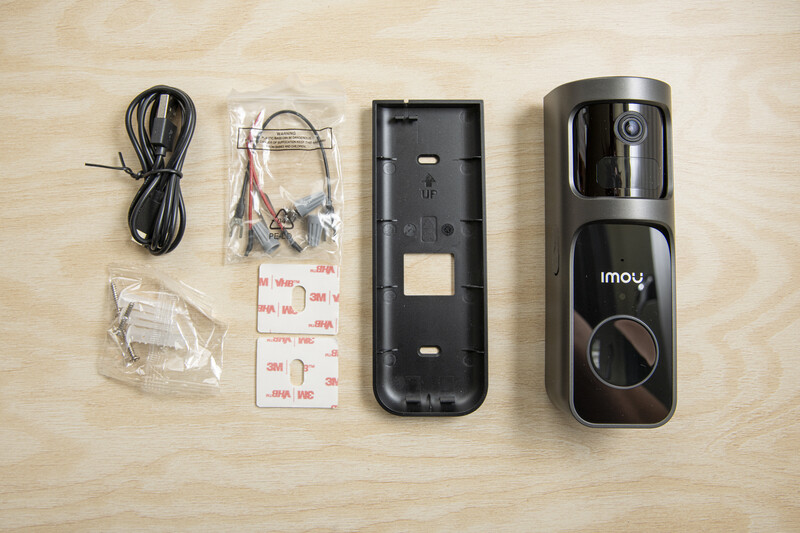 Imou smart video doorbell unboxing lay flat, displaying camera unit, power cable, mounting bracket, and adhesive strips.
