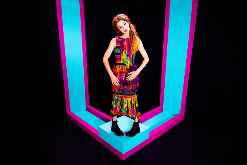 Woman in colorful fringed dress posing against neon cyan and magenta geometric backdrop.