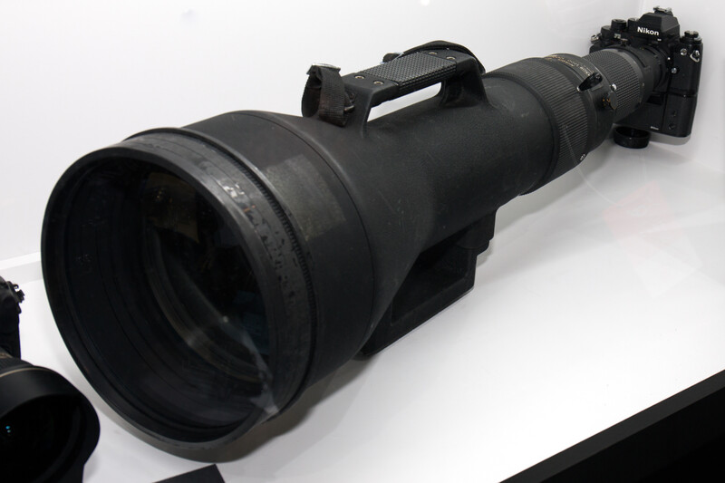 Large telephoto lens mounted on camera body with carrying handle, positioned horizontally on white surface.
