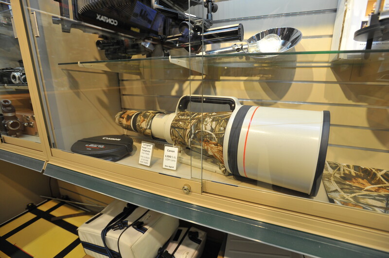 Telephoto lens with cream and black barrel displayed in a glass museum case.