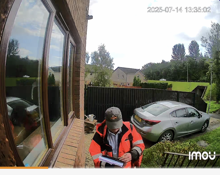 Person in orange safety vest examining a residential property exterior with doorway and parked silver sedan visible.