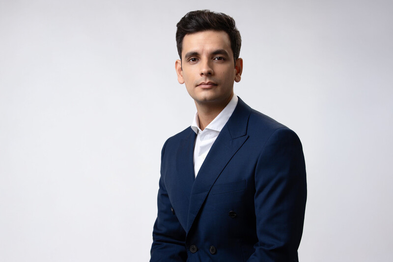 Professional headshot of a man in a dark navy double-breasted blazer and white dress shirt against a neutral gray background.
