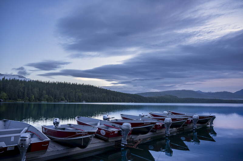 Row of moored boats on still water at dusk with forested shoreline and mountains under dramatic cloudy sky.