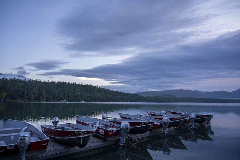 Moored wooden boats on still water at dusk with forested shoreline and mountains in background.