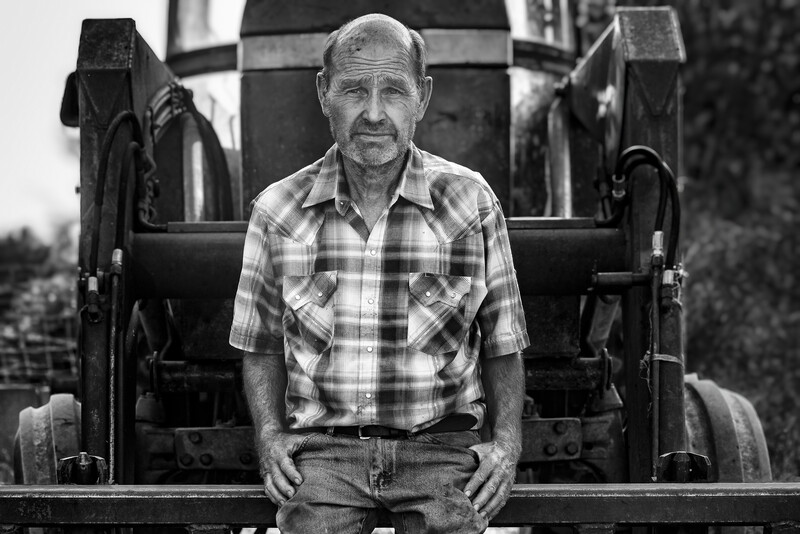 Man in plaid shirt sitting on vintage farm equipment, black and white portrait.